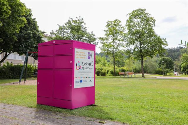 En rosa förvaringsenhet med flera fack står på en gräsmatta i en parkmiljö. På enheten finns en stor skylt med texten ”Leksaksbiblioteket” och illustrationer av leksaker. I bakgrunden syns gröna träd, en lekplats med gungställning och en gångväg. Himlen är ljus med moln.