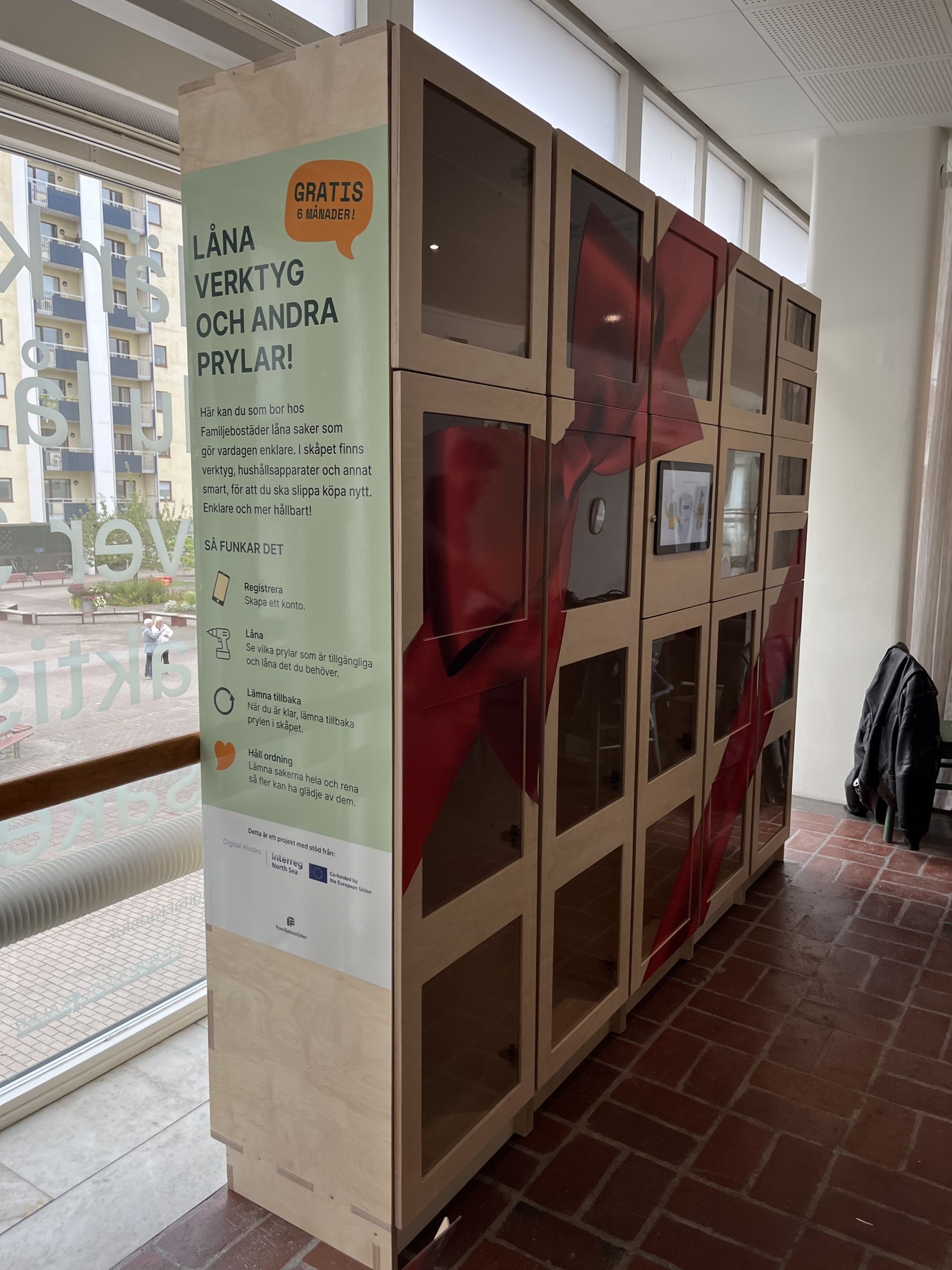 En modern, ljus lokal med smarta förvaringsskåp längs väggen. En person står framför skåpen och tittar på en digital skärm, medan en annan sitter på en bänk bredvid. Skåpen innehåller olika föremål och några är låsta. Växter och en grön matta ger en inbjudande känsla. Över skåpen finns tre informationsskyltar med instruktioner.