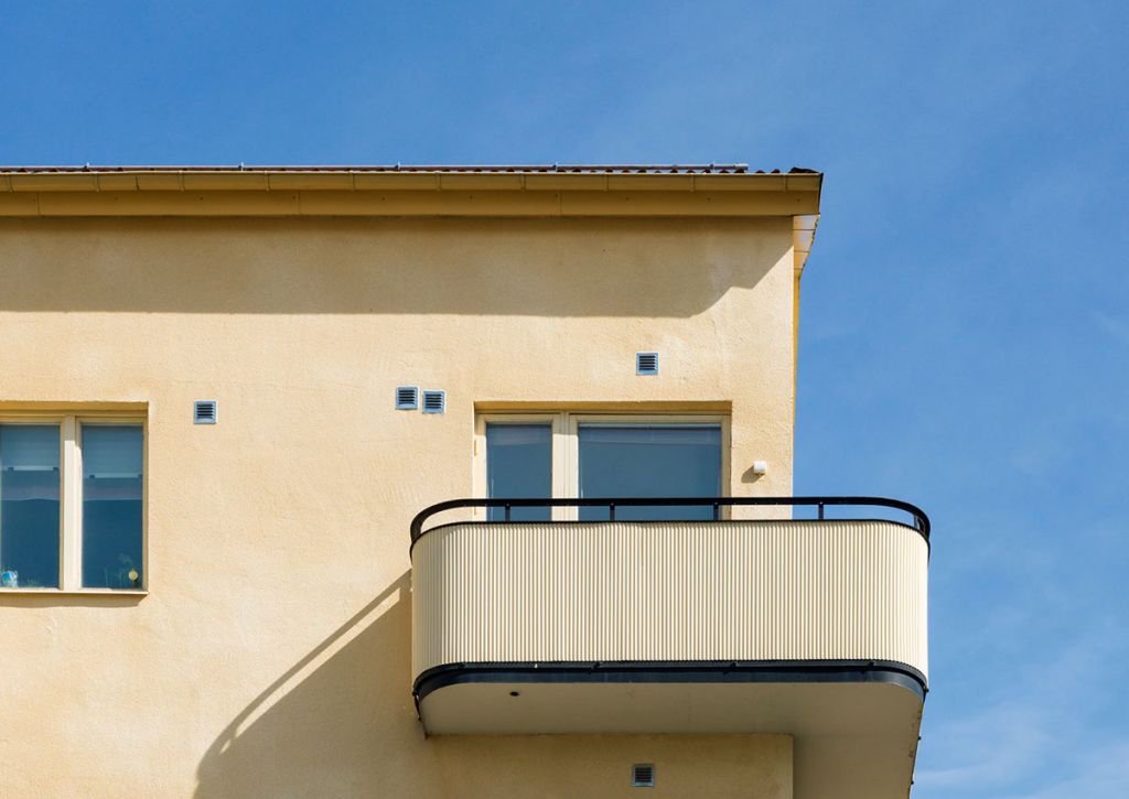 En rundad balkong som sträcker sig över två sidor av ett ljust gult hus med blå himmel i bakgrunden huset.